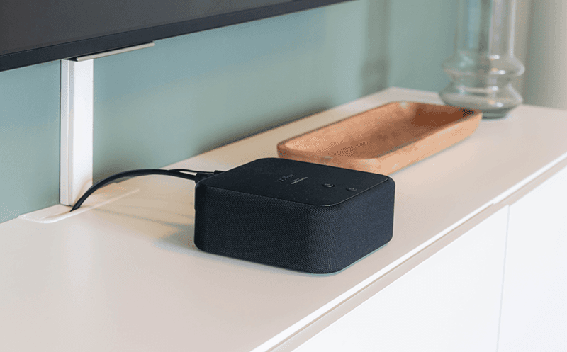 Sort YouSee Audio‑boks placeret på et hvidt tv‑møbel under et tv, med strømkabel og HDMI-stik tilsluttet bagpå.
