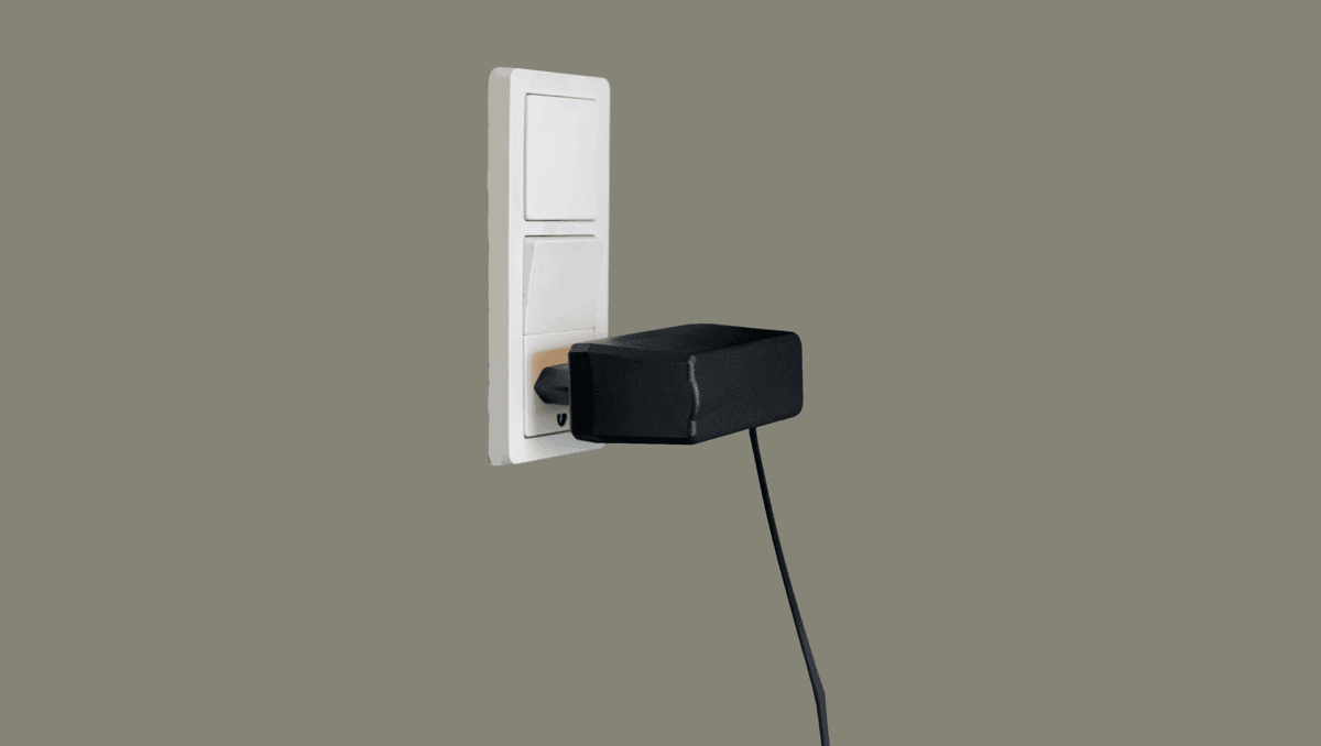 Sort strømadapter med tilsluttet kabel sat i hvid stikkontakt, som er tændt, vist på neutral baggrund.